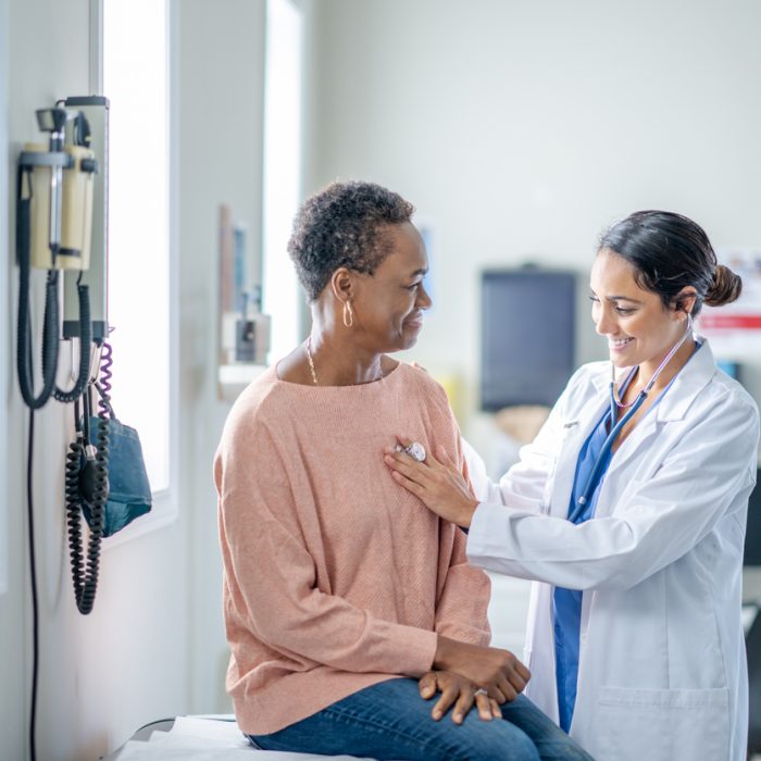 A senior woman of African decent sits up on an exam table during a routine check-up. She is sitting still and quiet as her female doctor listens to her heart.