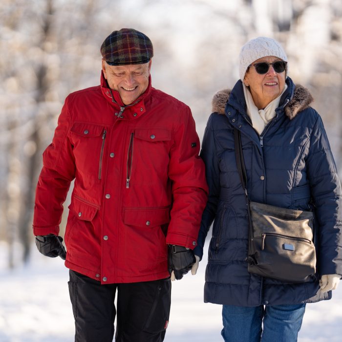 happy senior couple walking hand in hand on wide path of city public park lined with snow covered trees on cold sunny winter day, shallow focus on people, background blurred