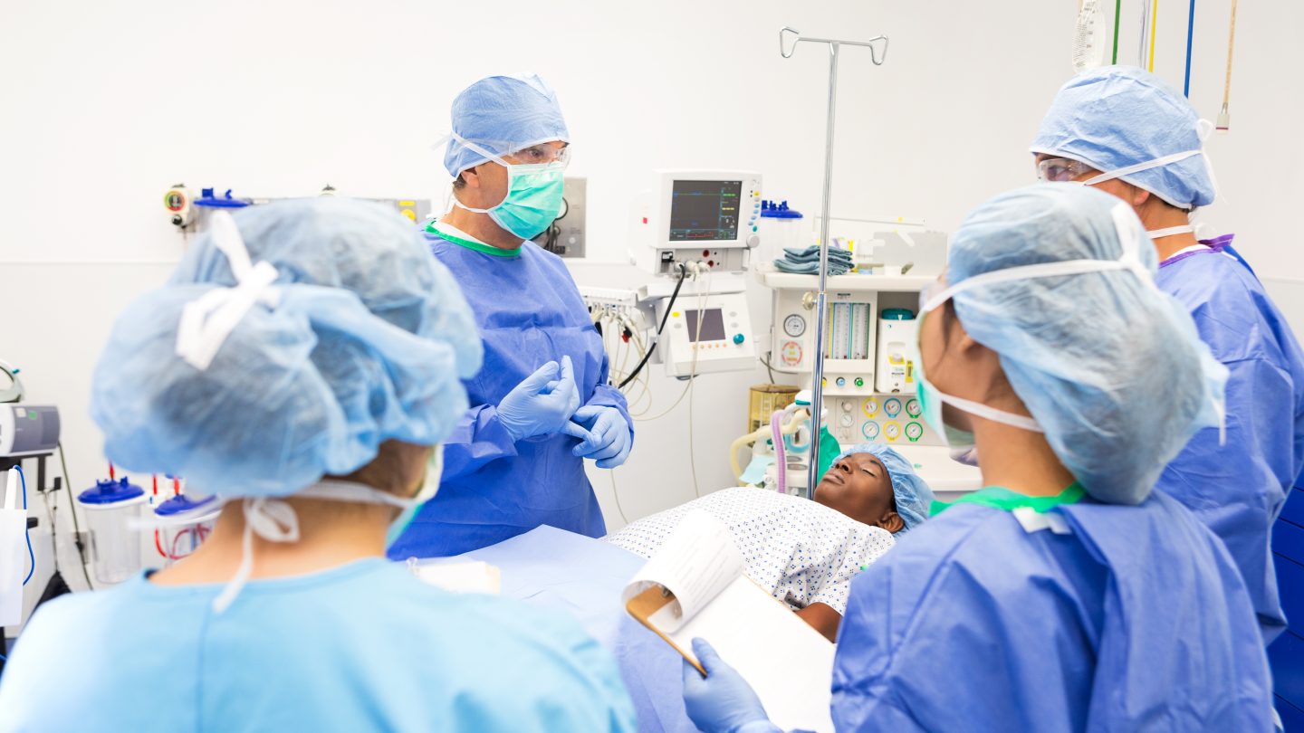 Lead surgeon is explaining surgical procedure to interns in teaching hospital while prepping patient for surgery. Doctors are wearing sterile blue surgical gowns, caps, masks, and gloves. Patient is lying on operating table.