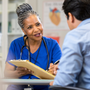 A senior female nurse sits at a table across from a new patient. She reaches across the table with a clipboard and points to the paperwork with a pen.