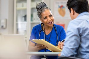 A senior female nurse sits at a table across from a new patient. She reaches across the table with a clipboard and points to the paperwork with a pen.
